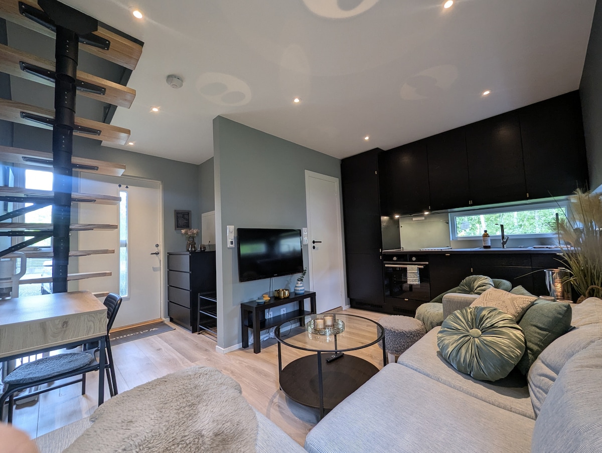 An open-plan living area is featured, showcasing a comfortable sectional sofa with soft cushions. A modern dining table is positioned nearby. A staircase leads to the upper level, and a sleek kitchenette is visible, complemented by contemporary cabinetry and ample natural light from the windows.