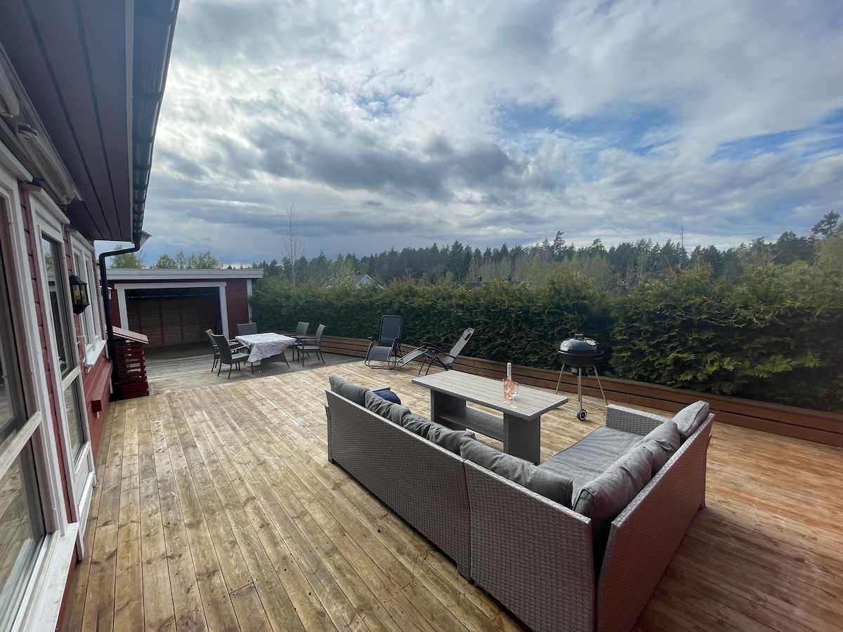 A spacious terrace is presented with wooden decking, featuring a cozy seating area with a sectional sofa and a central table. Several lounge chairs are arranged nearby, alongside a grill. Lush greenery outlines the perimeter, adding a sense of privacy to the outdoor space.