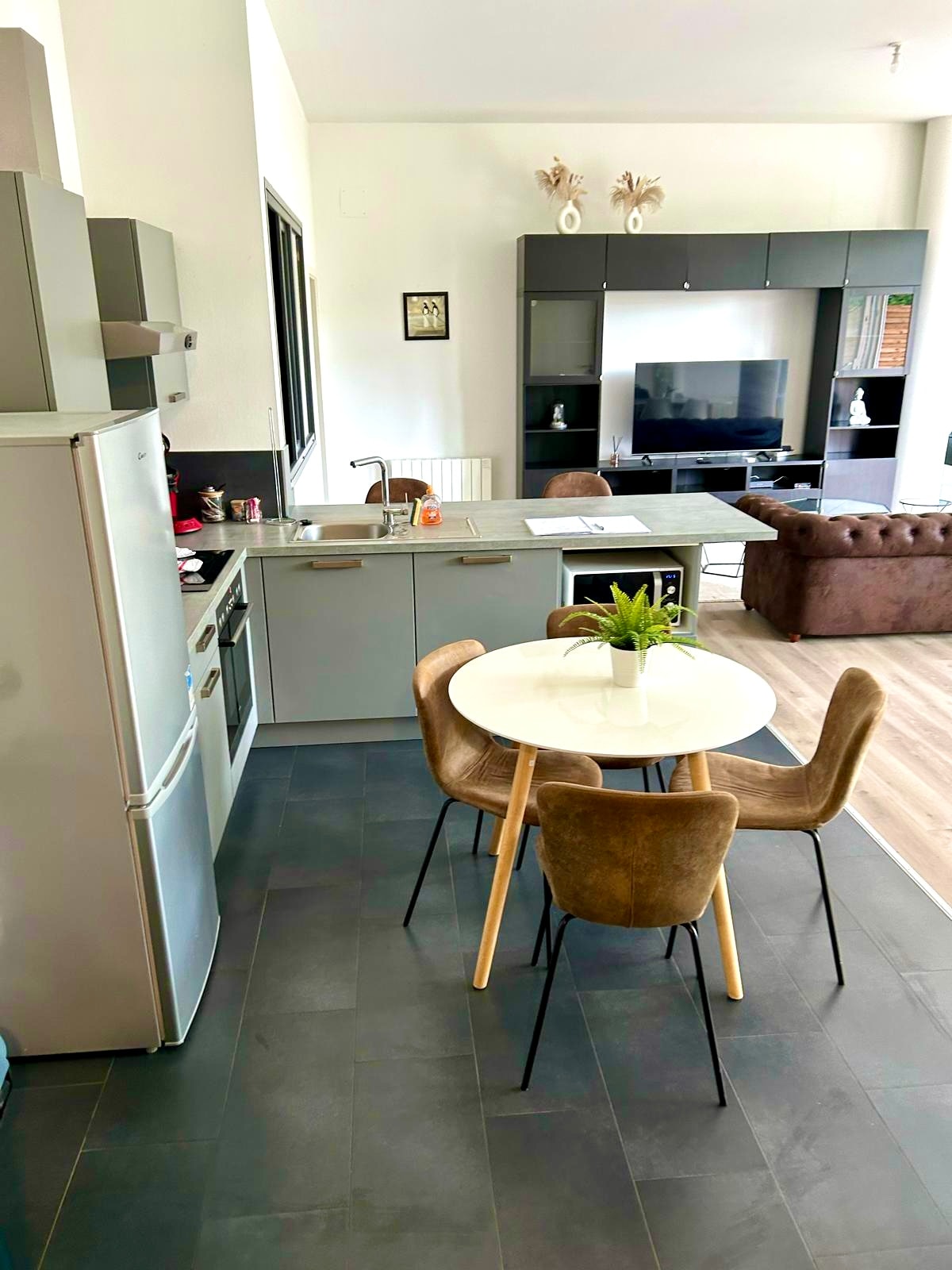 A modern kitchen area is visible, featuring sleek grey cabinetry and stainless steel appliances. A round dining table with four brown upholstered chairs is positioned in front of the counter. Natural light fills the space through a large window, illuminating the room's neutral tones.