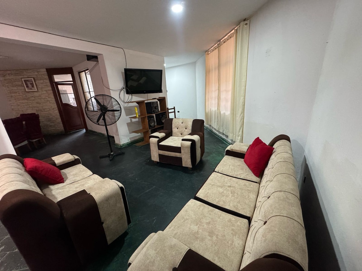 A spacious living area features comfortable seating, including a light-colored sofa set with accent pillows. A TV mounted on the wall is visible, with a floor fan positioned nearby. Natural light enters through a window adorned with sheer curtains, illuminating the open layout.