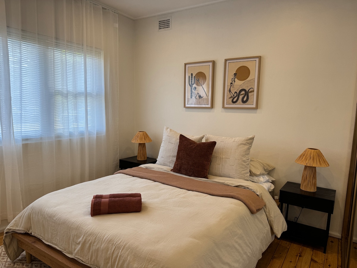 A well-appointed bedroom features a spacious bed adorned with neutral-toned linens and layered pillows. Two bedside lamps with soft, woven shades sit atop dark wood nightstands. Art prints are displayed above the bed, and a partially curtained window allows natural light to enter.