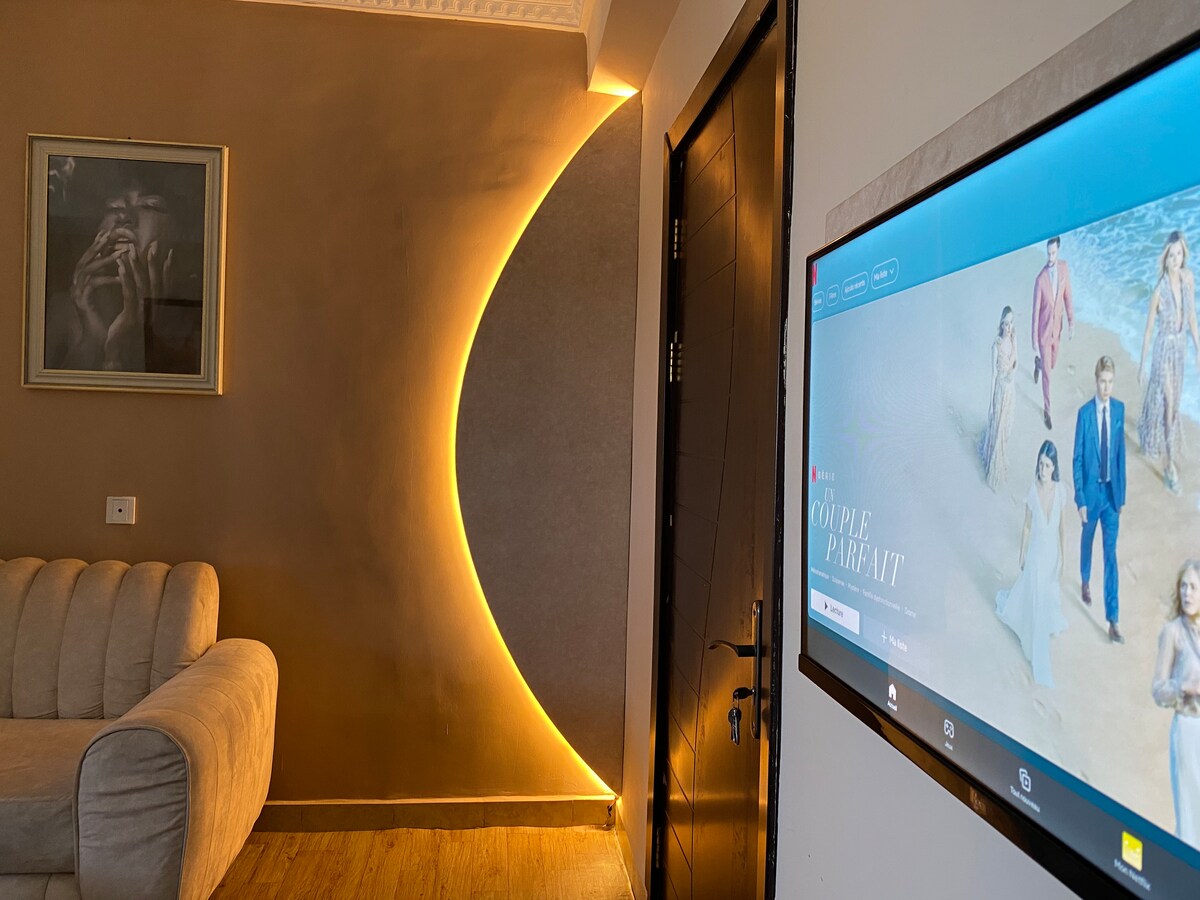 A cozy seating area features a modern beige sofa and a sleek television mounted on the wall. A soft glow from an elegantly shaped light fixture highlights the textured wall, adding a contemporary touch to the space. A framed art piece is positioned above the sofa.