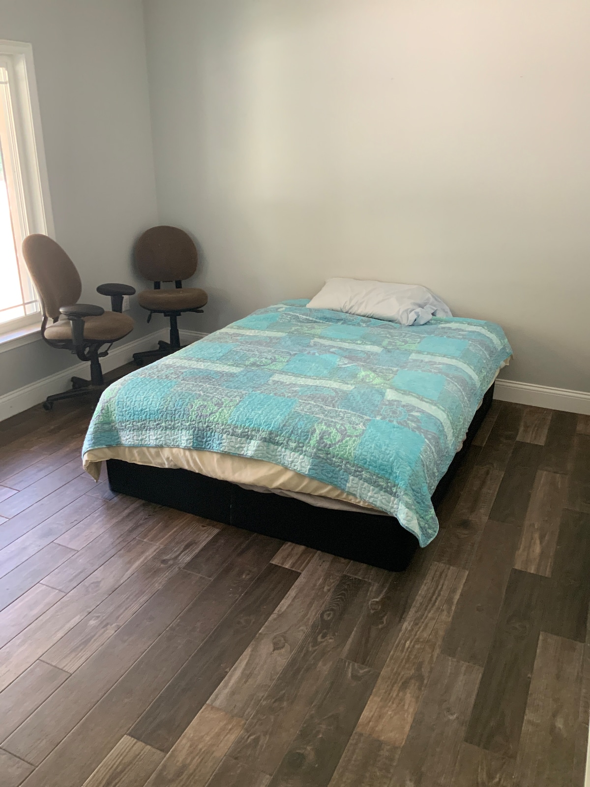 A spacious bedroom features a large bed with a light blue quilt and a white pillow. Two office chairs are positioned near a window, allowing natural light to illuminate the warm wooden floor.