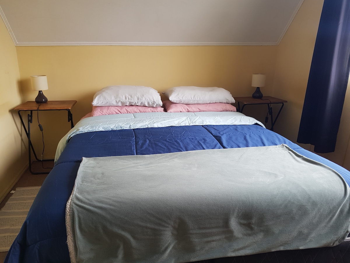 A spacious sleeping area features a large bed layered with soft bedding in pastel colors. Two bedside tables hold small lamps, and a textured rug lies underneath. Light filters in through a window dressed with dark curtains, complementing the warm yellow walls.