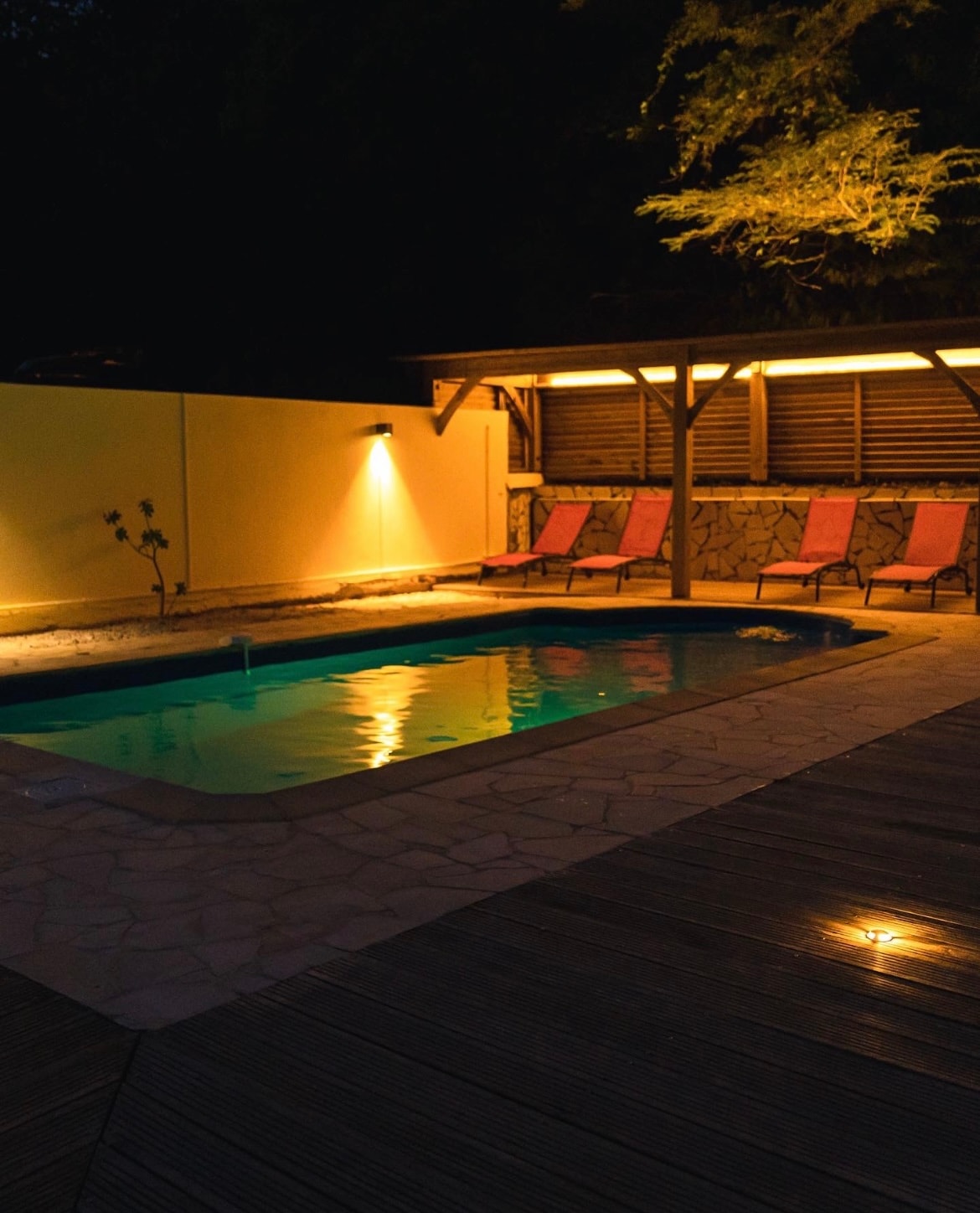 A serene pool area is illuminated by soft lighting, highlighting the tranquil water surface. Lounge chairs with pink cushions are arranged nearby, positioned on a softly textured deck. The surrounding walls are painted a soothing hue, enhancing the peaceful ambiance of the space.