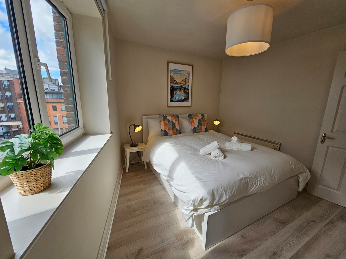 A cozy bedroom is highlighted by a spacious bed adorned with hotel-style linen and decorative pillows. Natural light fills the room through a large window, accompanied by a potted plant and bedside lamps, offering a gentle ambiance. A framed artwork is positioned above the bed.