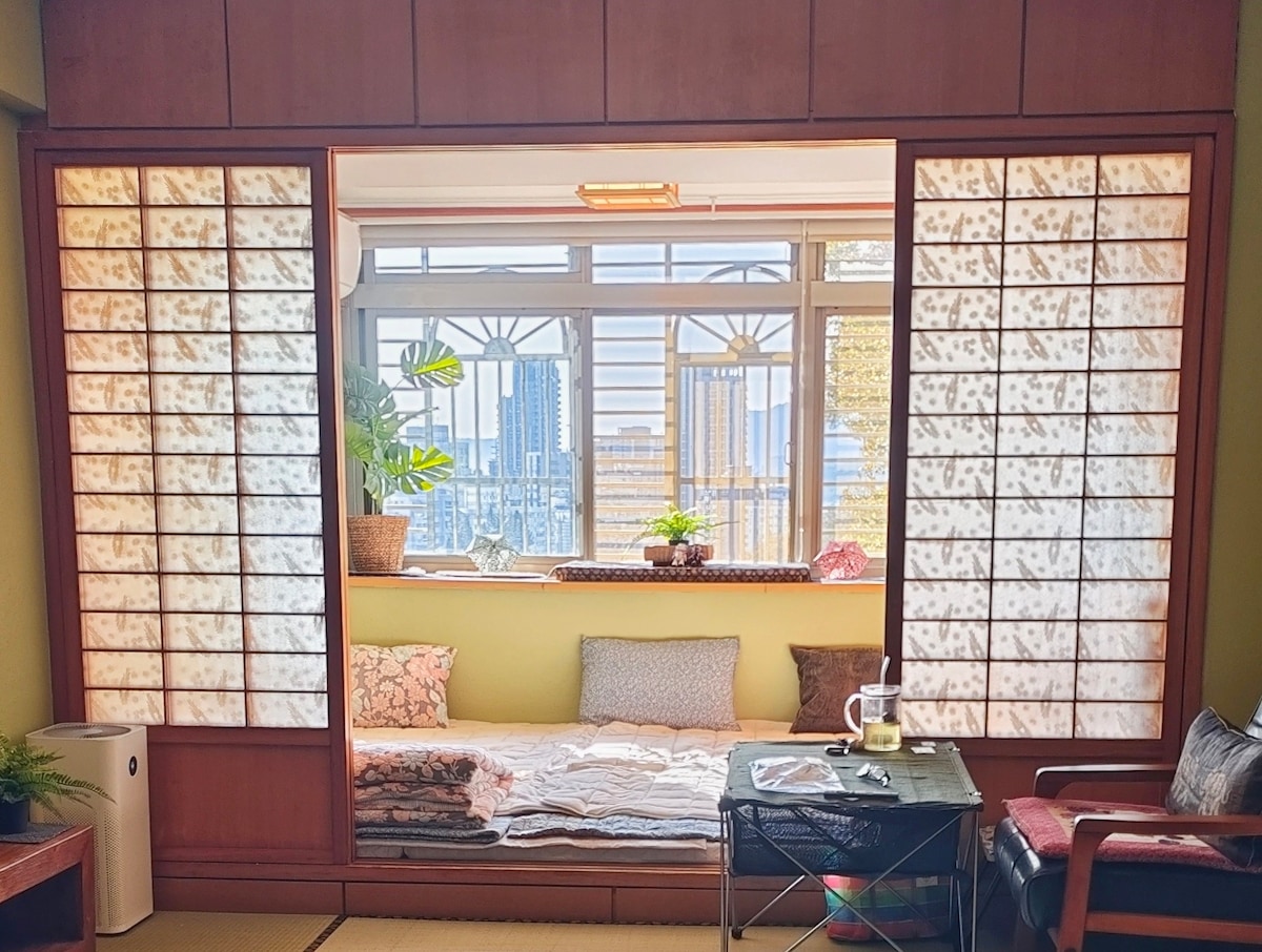 A spacious room features a traditional Japanese-style layout with sliding shoji doors and a cozy nook adorned with cushions. Potted plants are located on the windowsill, providing greenery and natural light. The view includes cityscape elements visible through large windows.