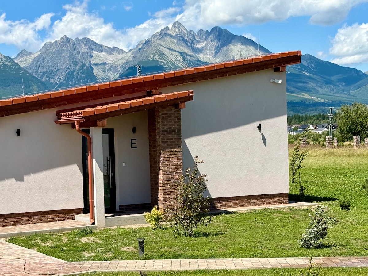 A modern building is shown with a distinctive roofline, featuring a welcoming entrance framed by manicured greenery. In the background, a mountain range extends under a clear blue sky, contributing to a serene landscape.