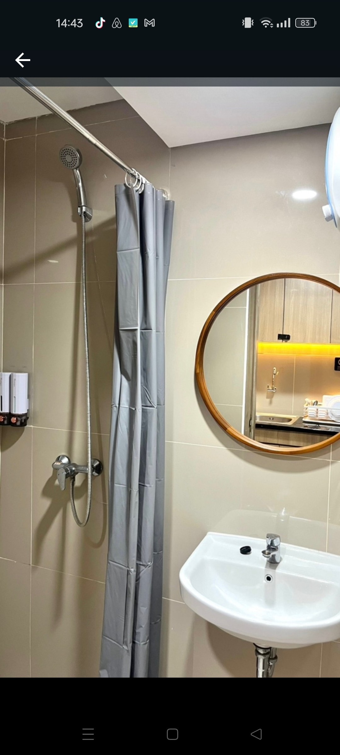 A clean bathroom area is showcased, featuring a round mirror above a compact sink. A gray shower curtain is seen beside a showerhead mounted on the wall. Neutral tiles create a simple yet functional space.