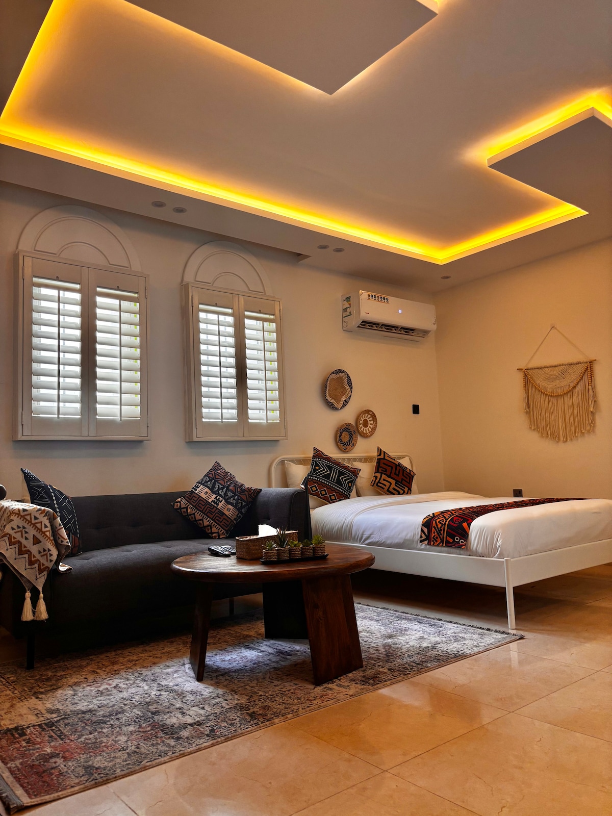 A serene bedroom area features a spacious bed with decorative pillows, complemented by a cozy gray sofa. Natural light enters through two shuttered windows, illuminating inviting wall decor. A round wooden table is positioned nearby, enhancing the comfort of the space.