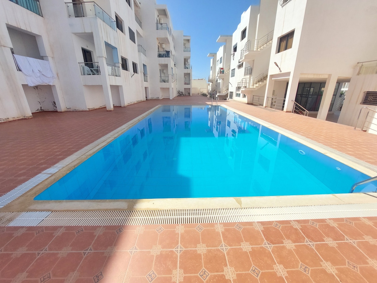 A spacious swimming pool is surrounded by modern residential buildings. The clear blue water reflects sunlight, enhancing the inviting atmosphere. Balconies with railings line the upper levels, while tiled flooring extends around the pool area, providing ample space for relaxation.