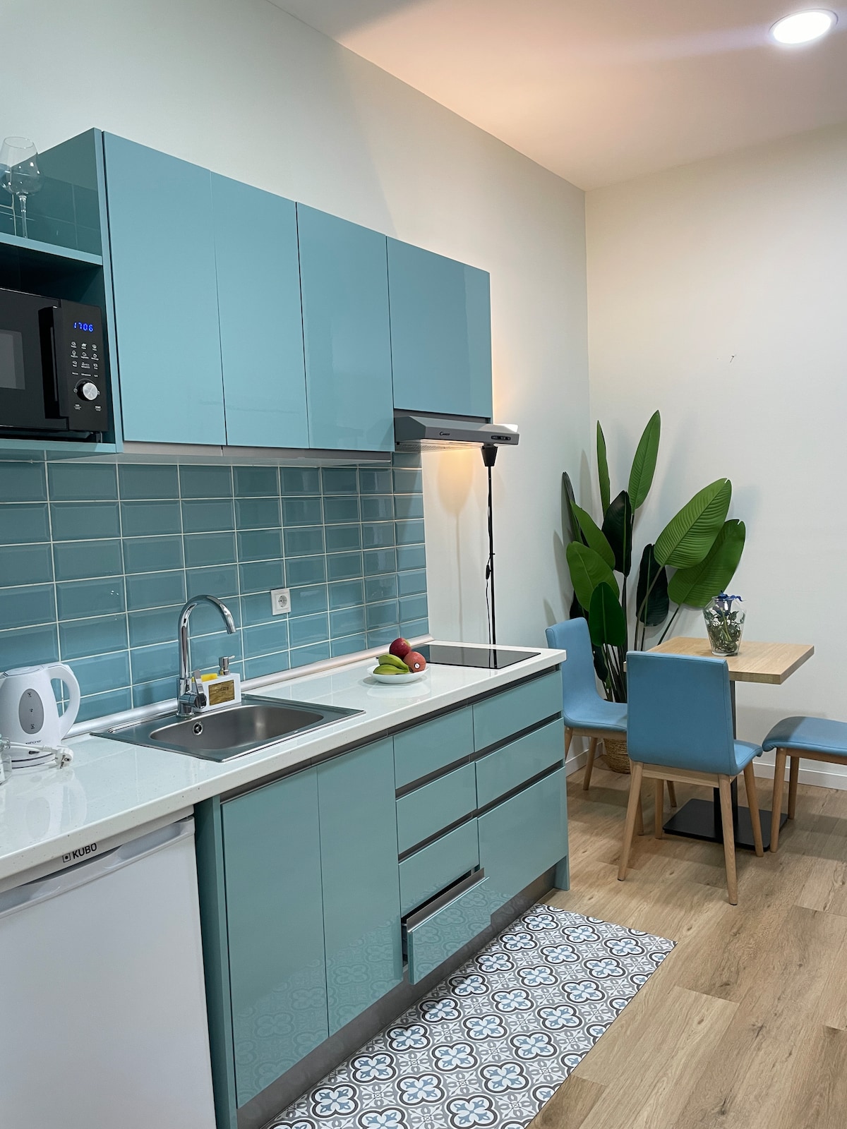 A modern kitchen is equipped with turquoise cabinetry and a white countertop. An integrated sink and microwave are visible. A small dining table with blue chairs is positioned nearby, accompanied by decorative plants, creating an inviting space for meal preparation and dining.