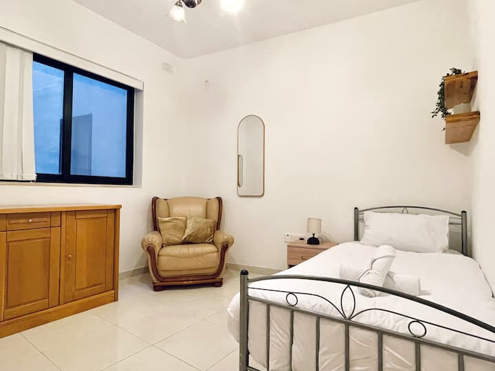 Deze lichte slaapkamer maximaliseert de ruimte met een passpiegel, een samenhangende inrichting en een uiterst comfortabele fauteuil bij het raam. Een rustig toevluchtsoord dat aanvoelt als een persoonlijk toevluchtsoord binnen de rustige stroom van de woning.