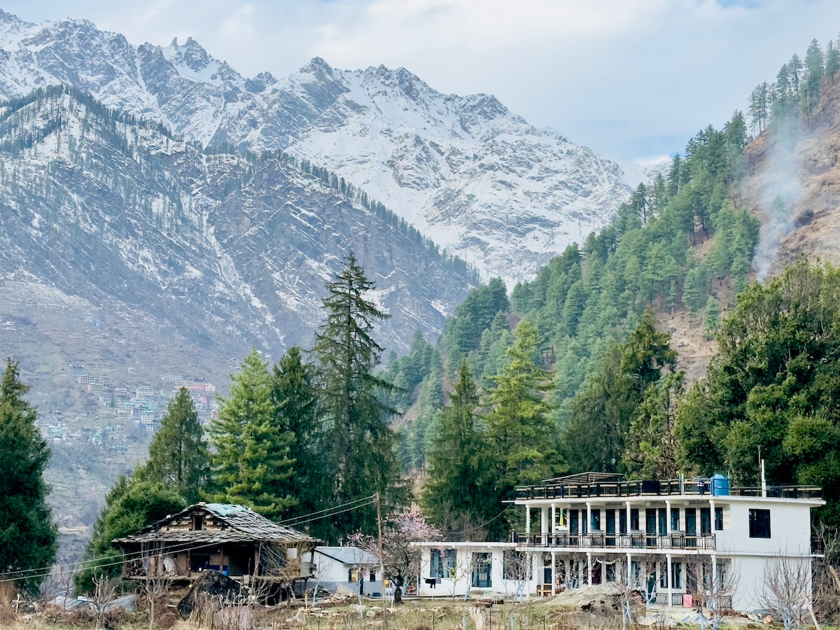 A guest house is nestled in a scenic mountain landscape, surrounded by tall trees. Snow-capped peaks rise in the background, while a rustic structure adds a charming element to the tranquil setting.