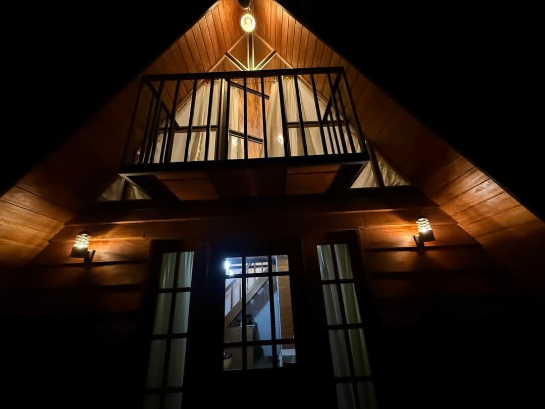 The cabin's exterior is illuminated at night, highlighting its wooden structure and sloped roof. A balcony with a railing is visible on the upper level, while warm lights illuminate the entryway below. Glass doors are framed by dark curtains, enhancing the cabin's inviting appearance.