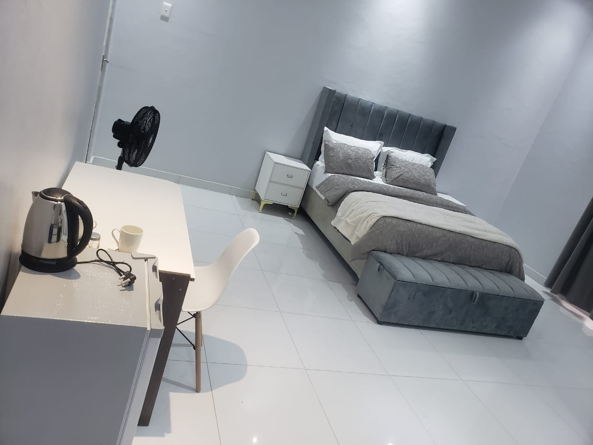 A spacious bedroom is visible, featuring a neatly made bed with plush pillows. A compact white desk accompanies a comfortable chair, complemented by a small kettle and cup. Soft gray tones dominate the decor, with smooth tile flooring and a floor fan adding to the space's functionality.