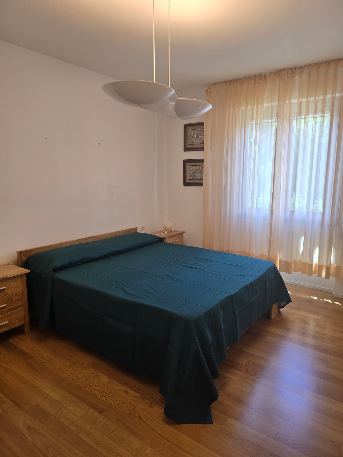 A serene bedroom features a spacious bed covered with a dark teal bedspread. Two bedside tables are positioned on either side, and large windows are dressed with light sheer curtains, allowing natural light to fill the room. Light wooden flooring complements the overall design.