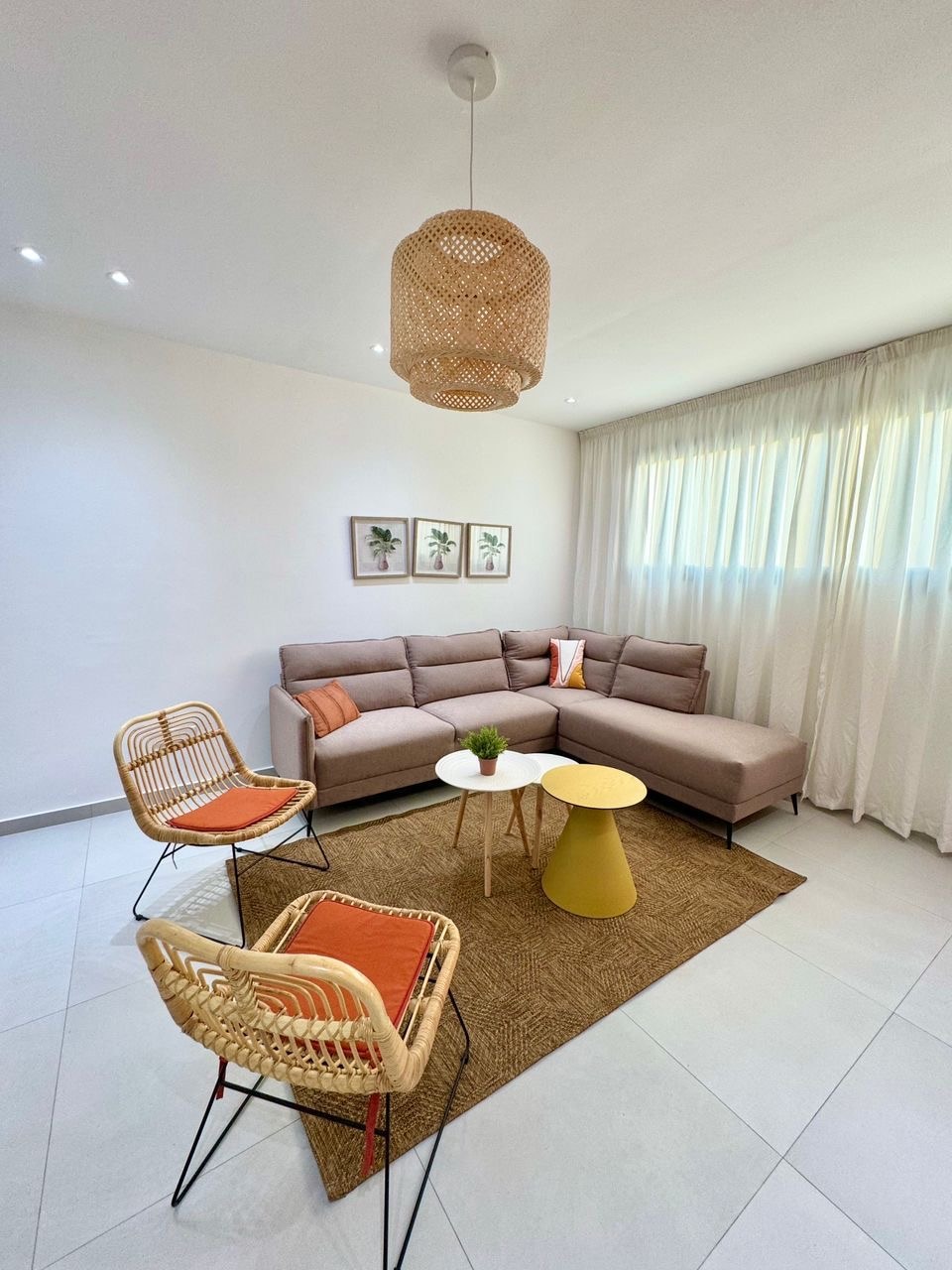 A modern living area is presented with a light-toned sectional sofa accompanied by two woven chairs and a round table. Soft, natural light filters through sheer curtains, enhancing the space. Framed botanical prints adorn the walls, adding an element of design to the minimalist décor.