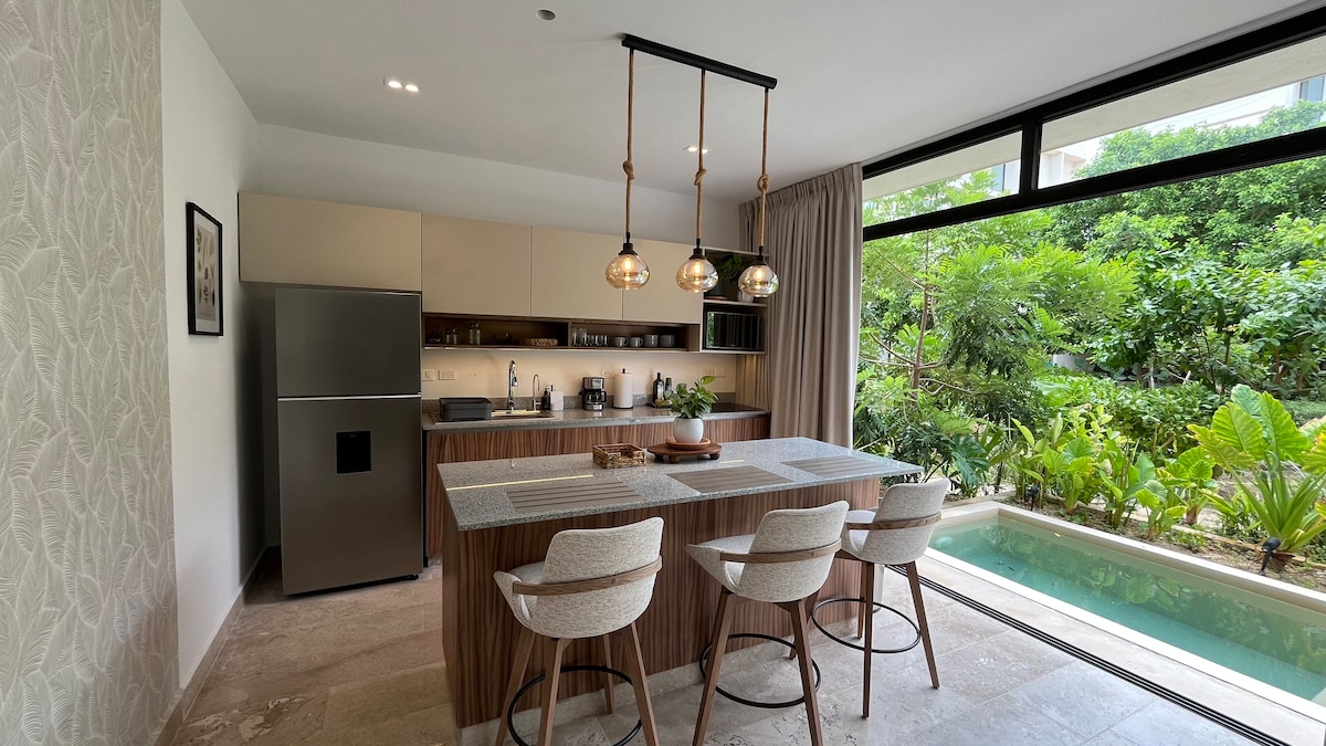 The modern kitchen showcases a spacious island with three counter stools and contemporary pendant lighting. Stainless steel appliances are integrated seamlessly, and large windows offer views of the lush greenery outside, enhancing the natural ambiance of the space.