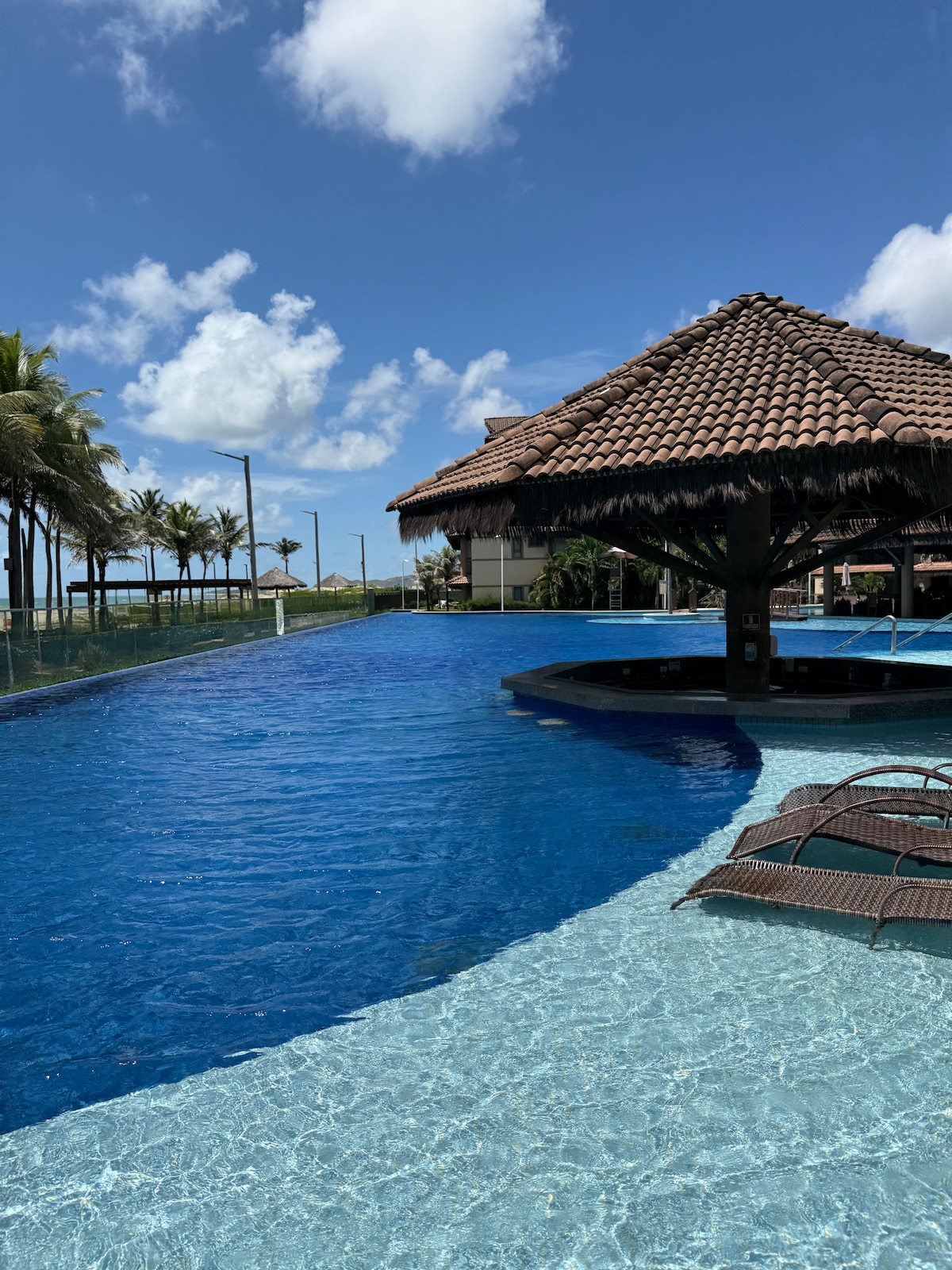 A glistening pool is designed with clear, turquoise water, surrounded by palm trees under a bright blue sky. A thatched-roof structure offers shade beside the water, while woven lounge chairs are positioned for relaxation near the pool's edge.