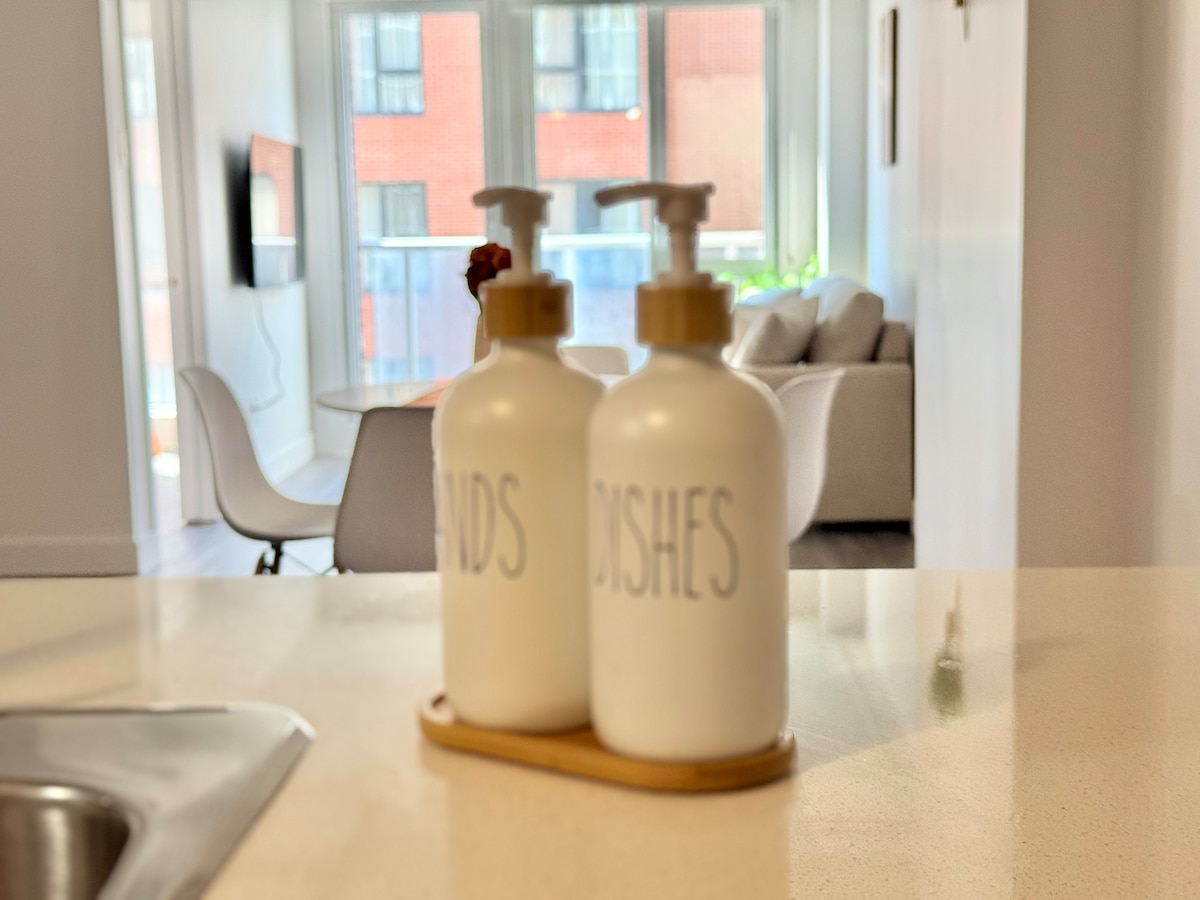 Two white pump bottles labeled 'HANDS' and 'DISHES' are positioned on a small wooden tray atop a countertop. In the background, a light-filled living area with a beige sofa is visible through large glass doors.