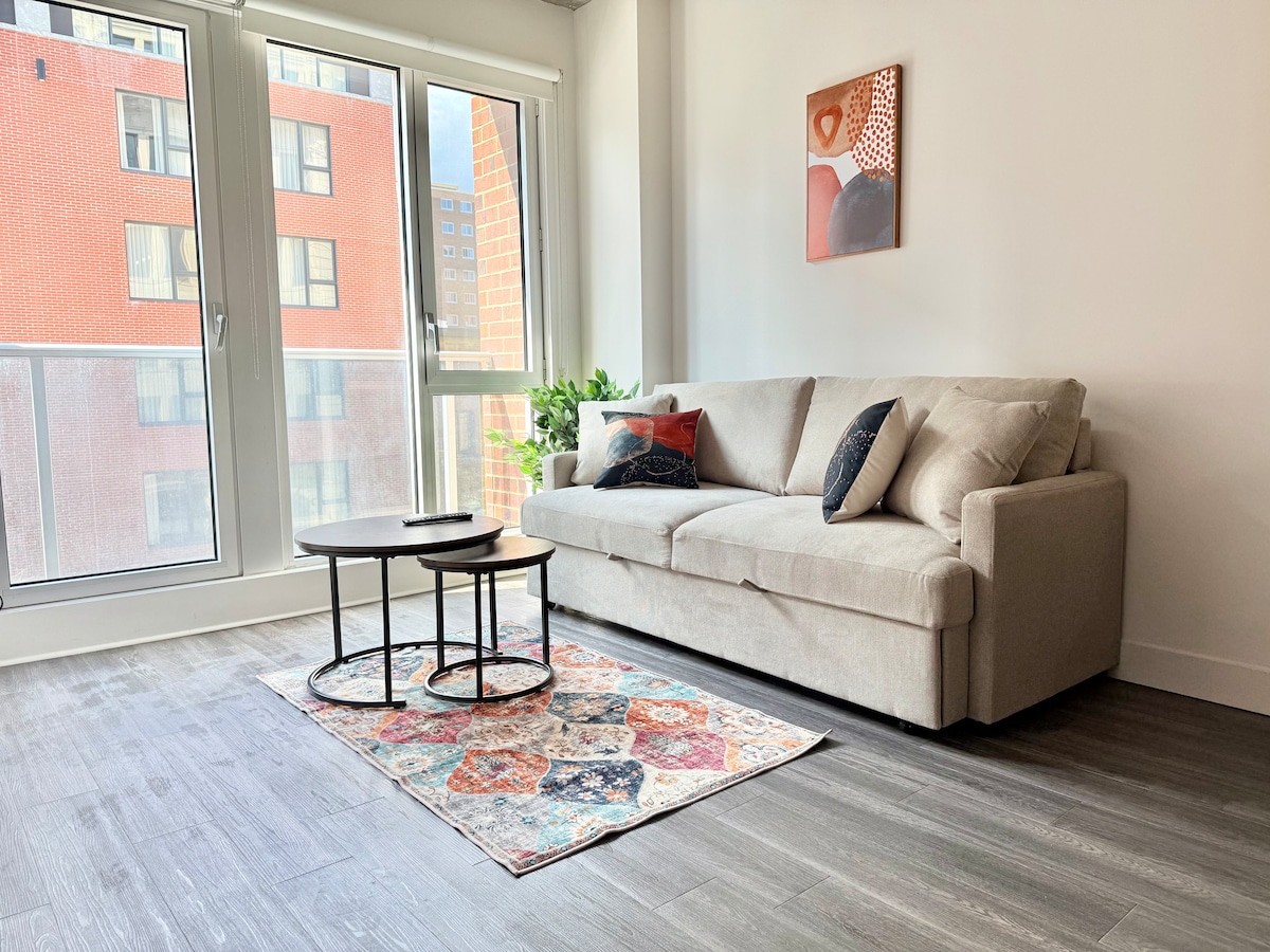 A well-lit living area features a light grey sofa with decorative cushions, positioned beside a large window. A small round table sits on a colorful area rug, which adds warmth to the space. Greenery from a nearby plant enhances the atmosphere.