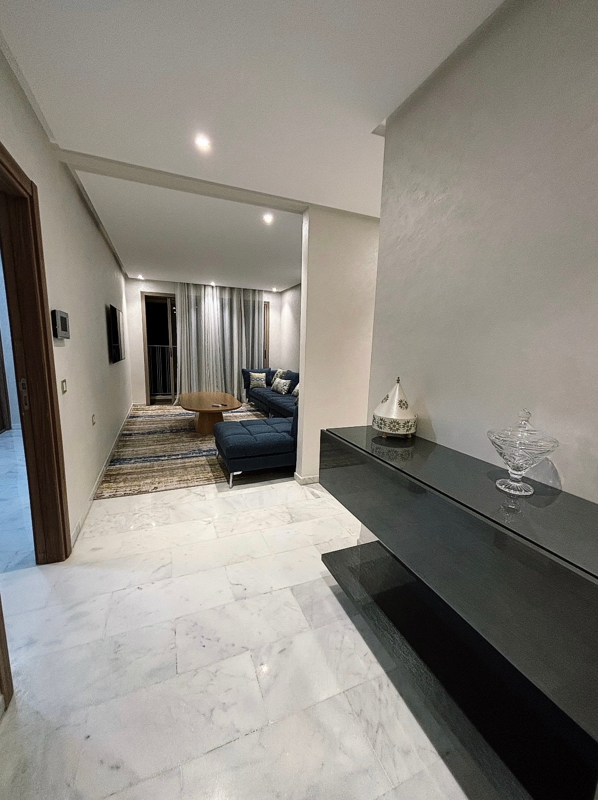A modern entryway is depicted, featuring a sleek black console table against a light-colored wall. A cozy seating area with a blue sofa and patterned rug can be seen in the background, illuminated by soft lighting. Large windows draped with light curtains allow for natural light.