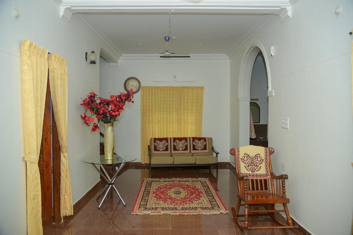 A spacious living area features a traditional design with a seating arrangement that includes a couch and a rocking chair. A decorative rug lies on the floor, complemented by a glass table with a floral arrangement. Soft yellow curtains frame the window, allowing natural light to illuminate the space.