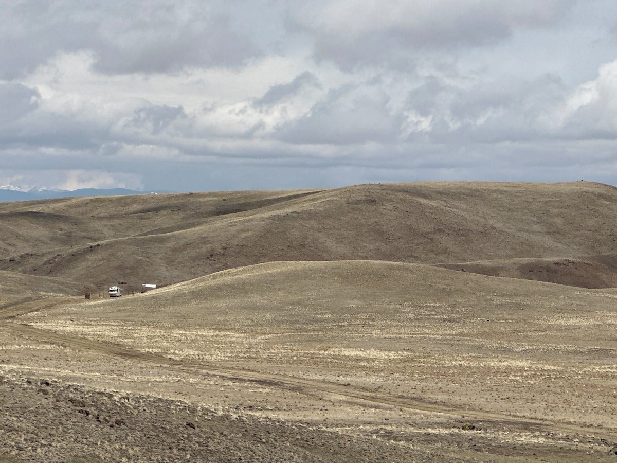 A vast open landscape is displayed with rolling hills covered in dry grasses. The expansive sky above showcases a mix of gray and white clouds, while a distant travel trailer is visible, situated along a winding dirt path.