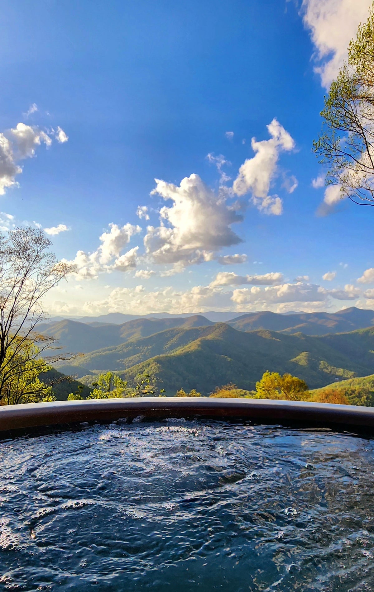 A hot tub sits at the edge of a deck, surrounded by unobstructed views of rolling mountains and a vibrant sky. Soft clouds drift over layers of green foliage, enhancing the tranquil atmosphere of the scene.