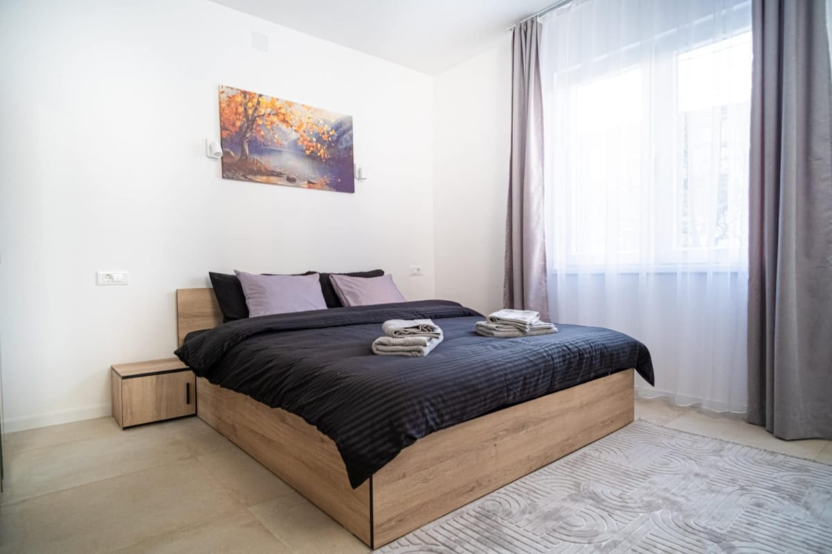 A modern sleeping area features a spacious bed with dark bedding, complemented by light purple pillows. A small nightstand rests beside the bed. Natural light enters through a large window, which is adorned with sheer curtains, illuminating the light-toned walls and flooring.
