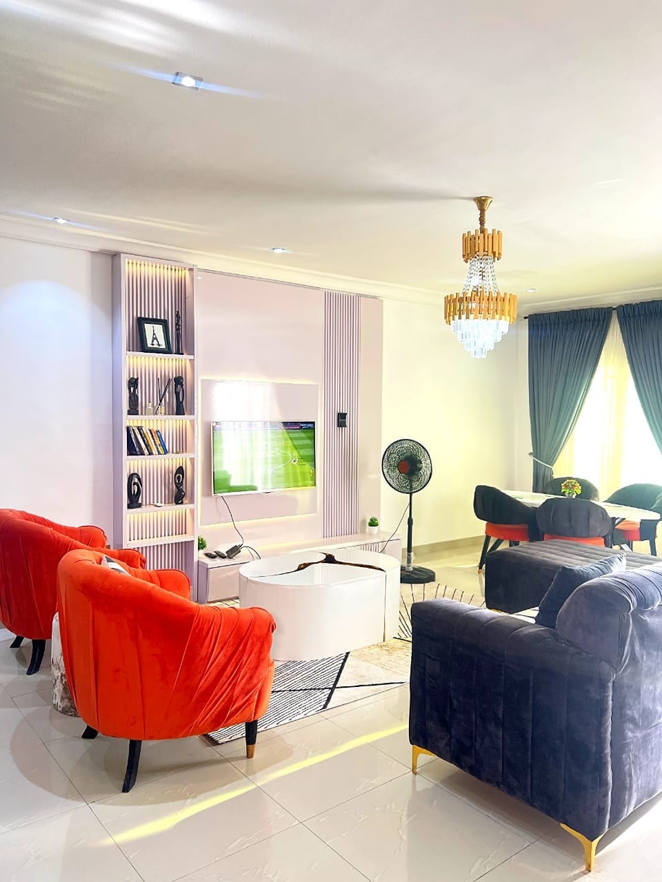 A spacious living area is showcased with contrasting seating options in vibrant orange and deep blue. A flat-screen TV is mounted on a wall, accompanied by shelves displaying decorative items. Large windows are draped with dark curtains, allowing abundant natural light to flow in.