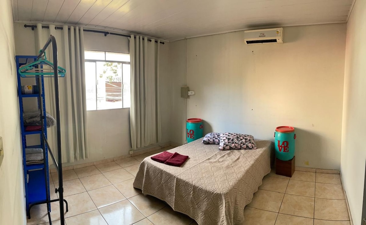 A spacious sleeping area is furnished with a large bed covered with a light quilt. Two colorful storage barrels are positioned at the foot of the bed. Natural light filters through a window adorned with light curtains, and a clothes rack stands in one corner.