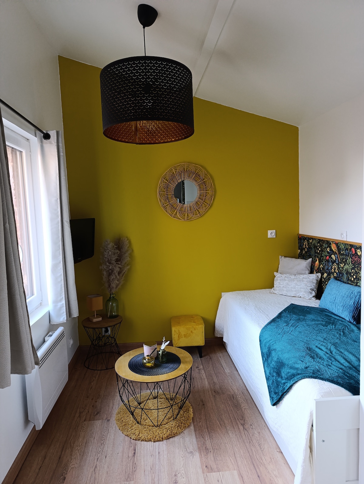 A bright sleeping area showcases a spacious bed adorned with soft linens. A decorative, vibrant wall accent adds color, while a small round table is paired with a textured yellow pouf. A mirror and a TV are present, enhancing the room's cozy atmosphere.