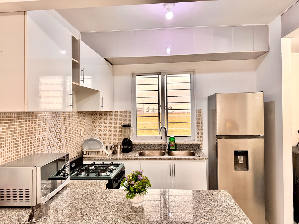 A modern kitchen features sleek white cabinetry and a mosaic-tiled backsplash. A granite countertop houses a gas stove and a stainless-steel refrigerator. Natural light fills the space through a large window, which frames a view of open fields.