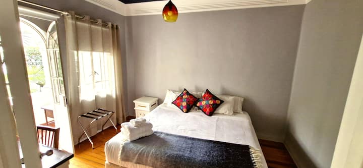 Habitación 4: con balcón propio al jardín interior, es la única habitación con vista abierta al exterior. Cama king size, mesas de noche blancas y cojines bordados en tonos joya.