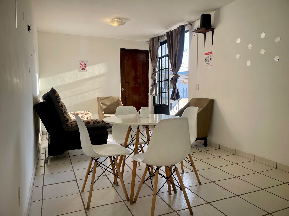 A cozy living area is presented with a circular dining table surrounded by four white chairs. A comfortable armchair and a larger sofa are visible, promoting relaxation. Natural light enters through a large window with light curtains, and the floor is tiled in light colors.