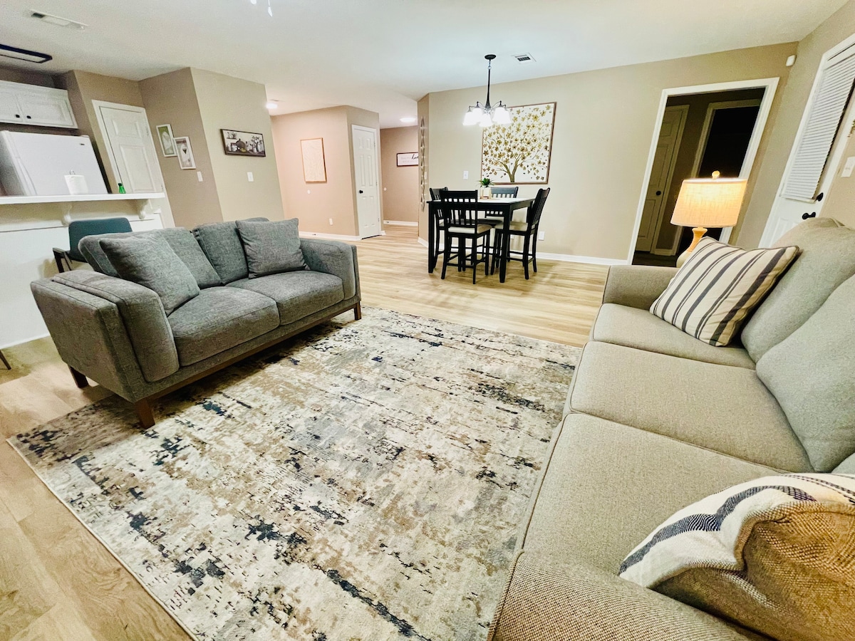 A contemporary living area features a comfortable gray sofa and a beige couch, positioned on a patterned area rug. A dining table with chairs stands in the background, illuminated by a stylish chandelier. Light wood flooring enhances the open layout of the space.