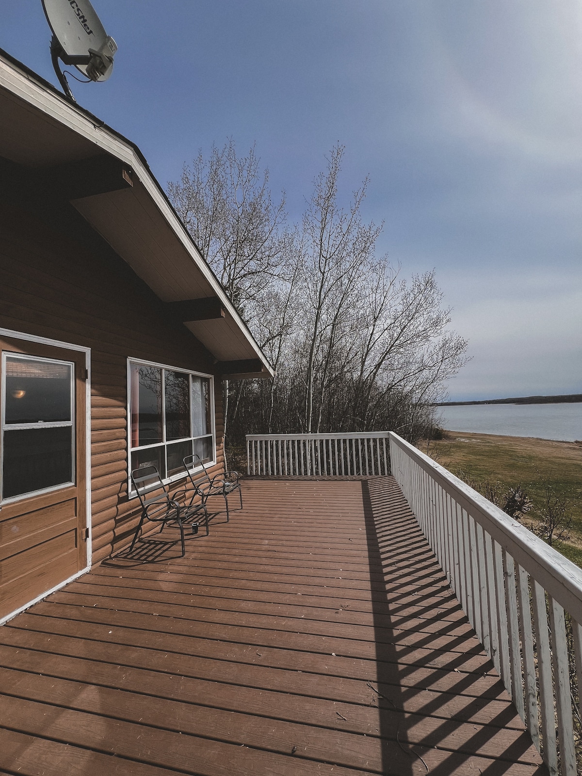 A spacious lakeside deck features wooden flooring and a white railing, providing a peaceful outdoor space. Sunlight reflects off the adjacent water, while trees line the edge of the property. A few chairs are positioned for enjoying the view.