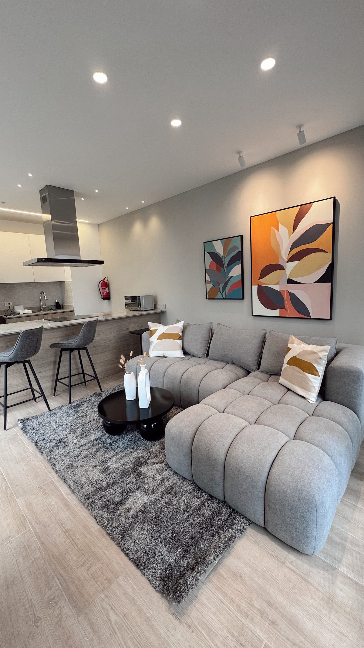 A modern living area features a gray sectional sofa adorned with decorative throw pillows. A round black coffee table sits on a soft area rug, and two bar stools are positioned at a kitchen counter in the background. Colorful artwork enhances the walls.