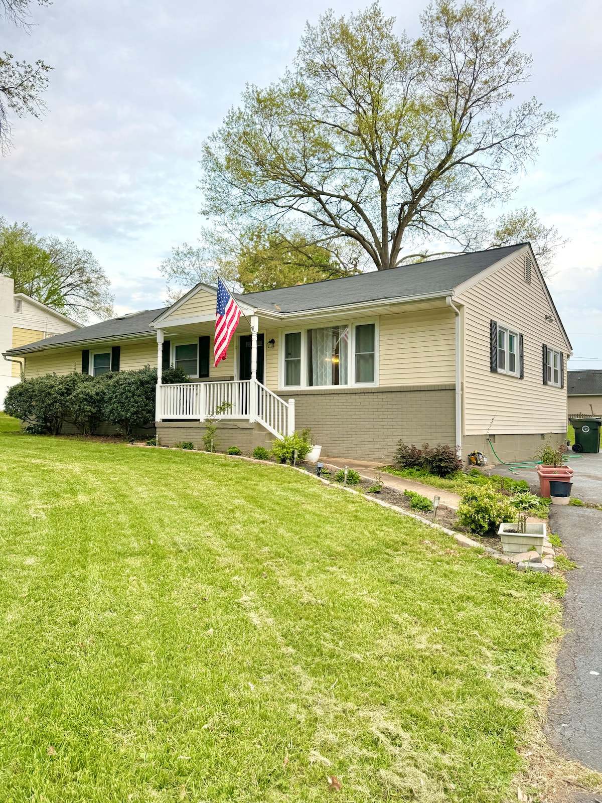 A single-story home is showcased, featuring a welcoming front porch with white railings and an American flag. Lush green grass and decorative plants line the pathway, while a large tree provides shade, enhancing the inviting exterior of the property.