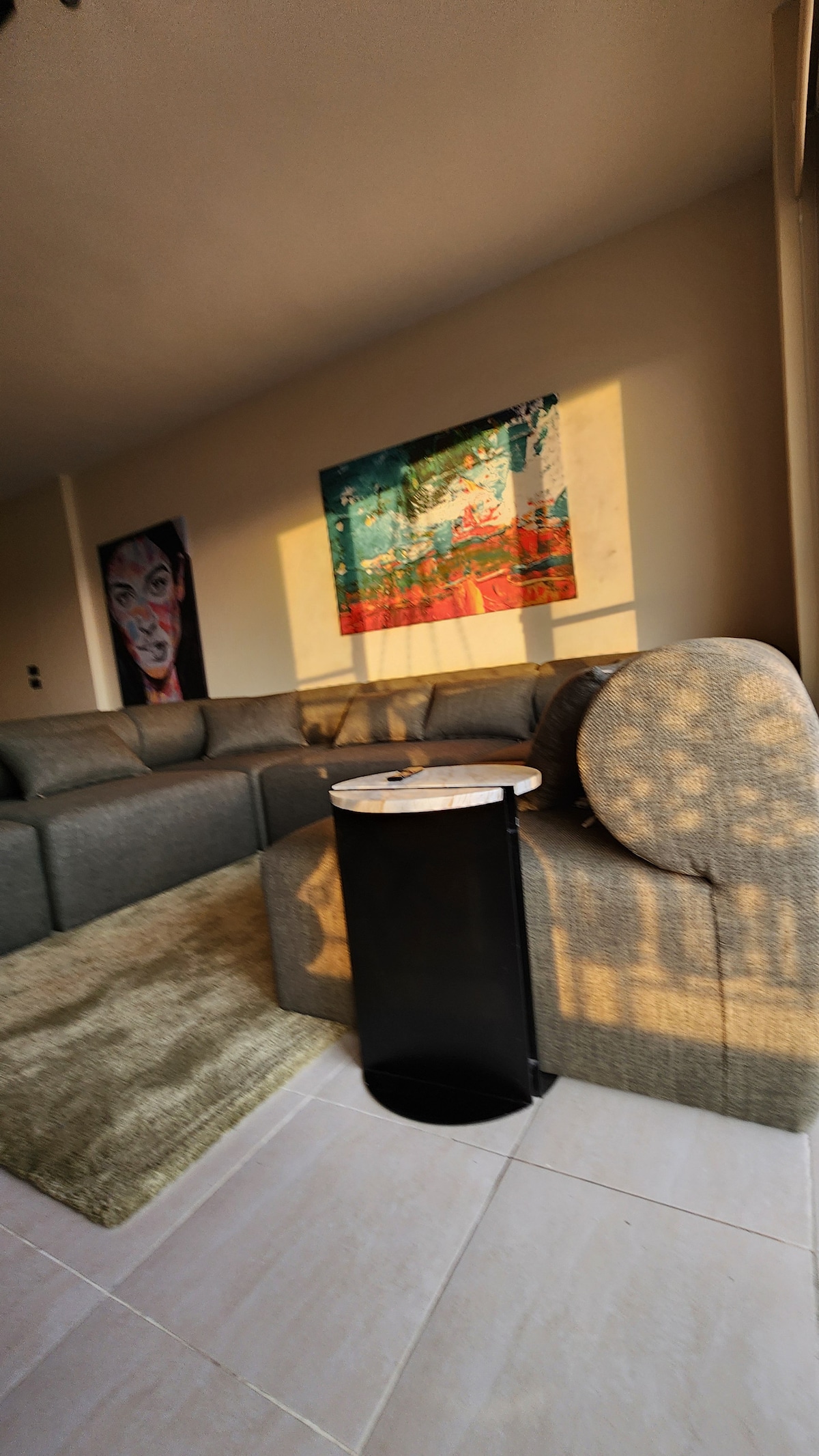 A modern living area is depicted, featuring a spacious gray sofa with an artistic wall hanging above it. A round side table rests beside the sofa, and natural light falls across the scene, highlighting the soft texture of an area rug beneath.