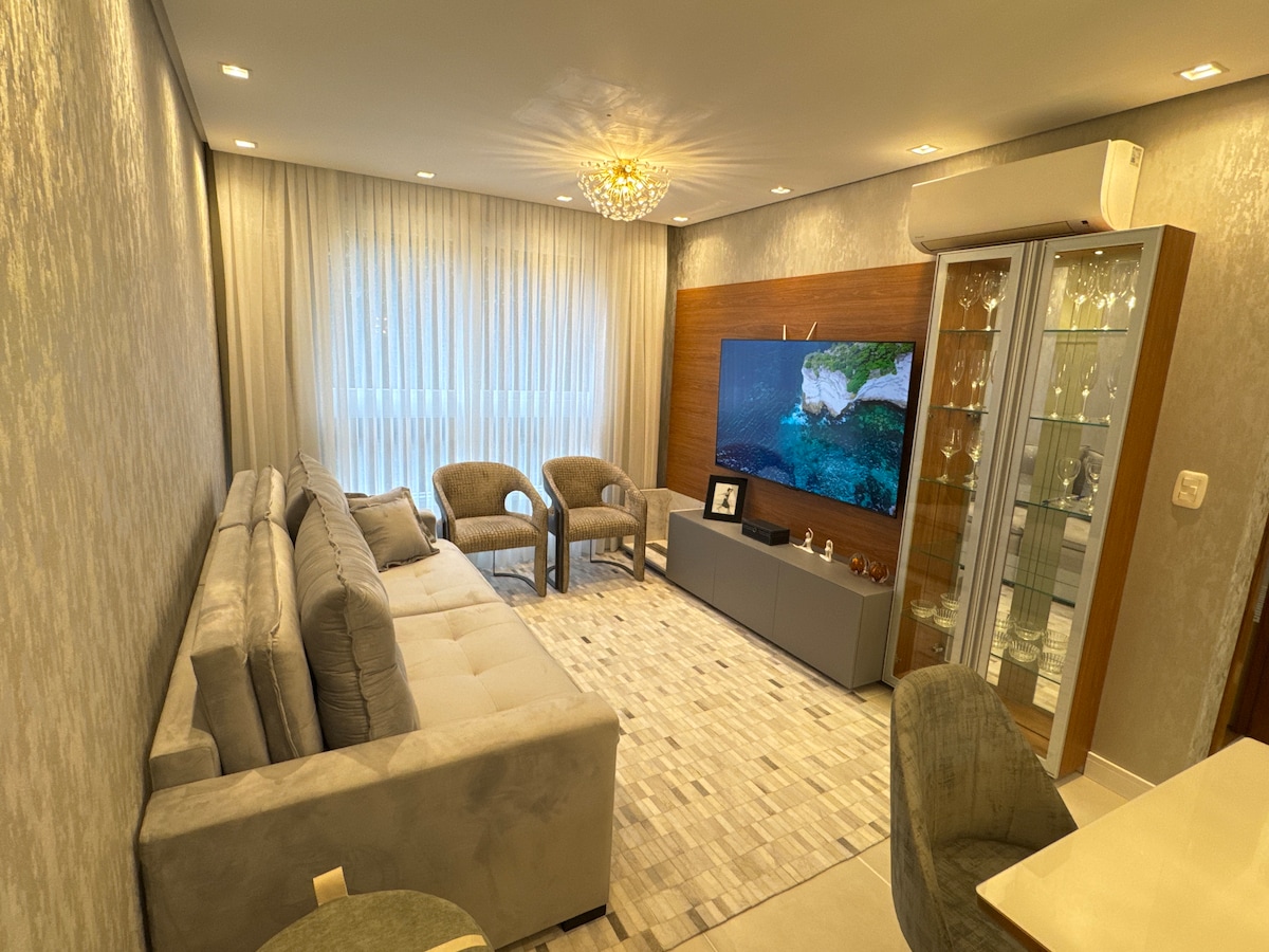 A modern living area is displayed, featuring a comfortable sofa and two armchairs arranged around a wide coffee table. A television is mounted on a wooden accent wall, while a glass display cabinet showcases decorative items. Soft natural light is filtered through sheer window curtains.