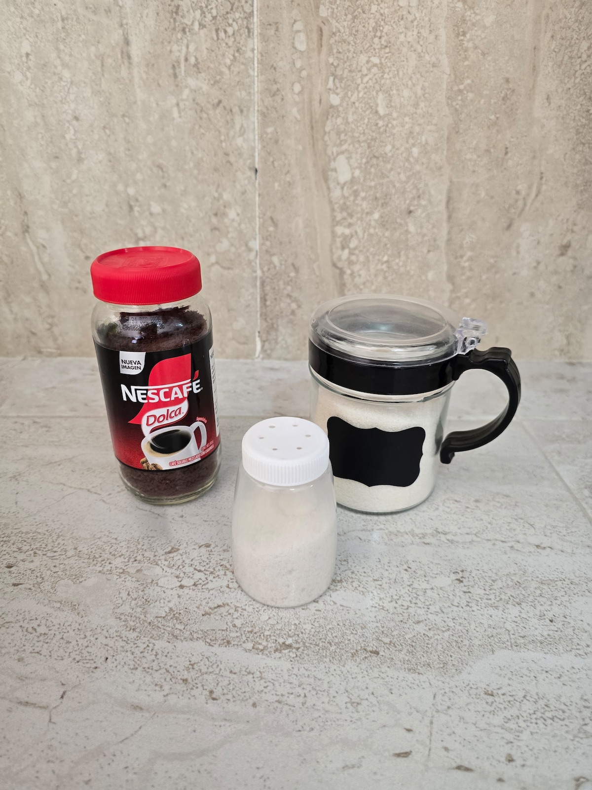 An arrangement of coffee essentials is presented on a neutral-toned surface. A jar of Nescafé coffee stands alongside a shaker of white powder and a clear mug with a black lid. The elements are placed for easy access, promoting a convenient coffee-making experience.