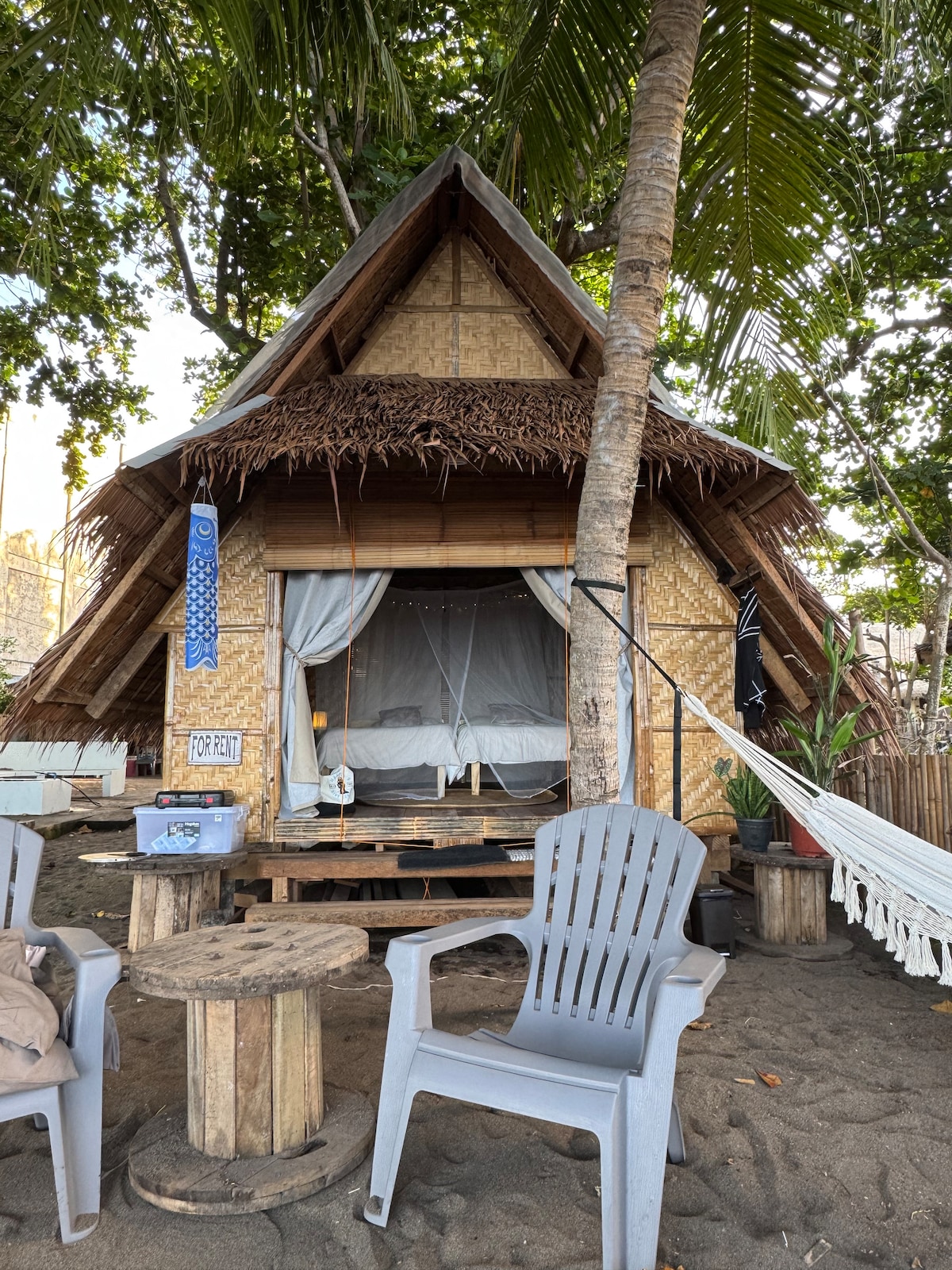 A charming nipa hut cottage stands at the ocean's edge, built with traditional materials and featuring a thatched roof. A covered sleeping area is visible, framed by light drapes. Outdoor seating consists of two gray chairs and rustic wooden tables on sandy ground.