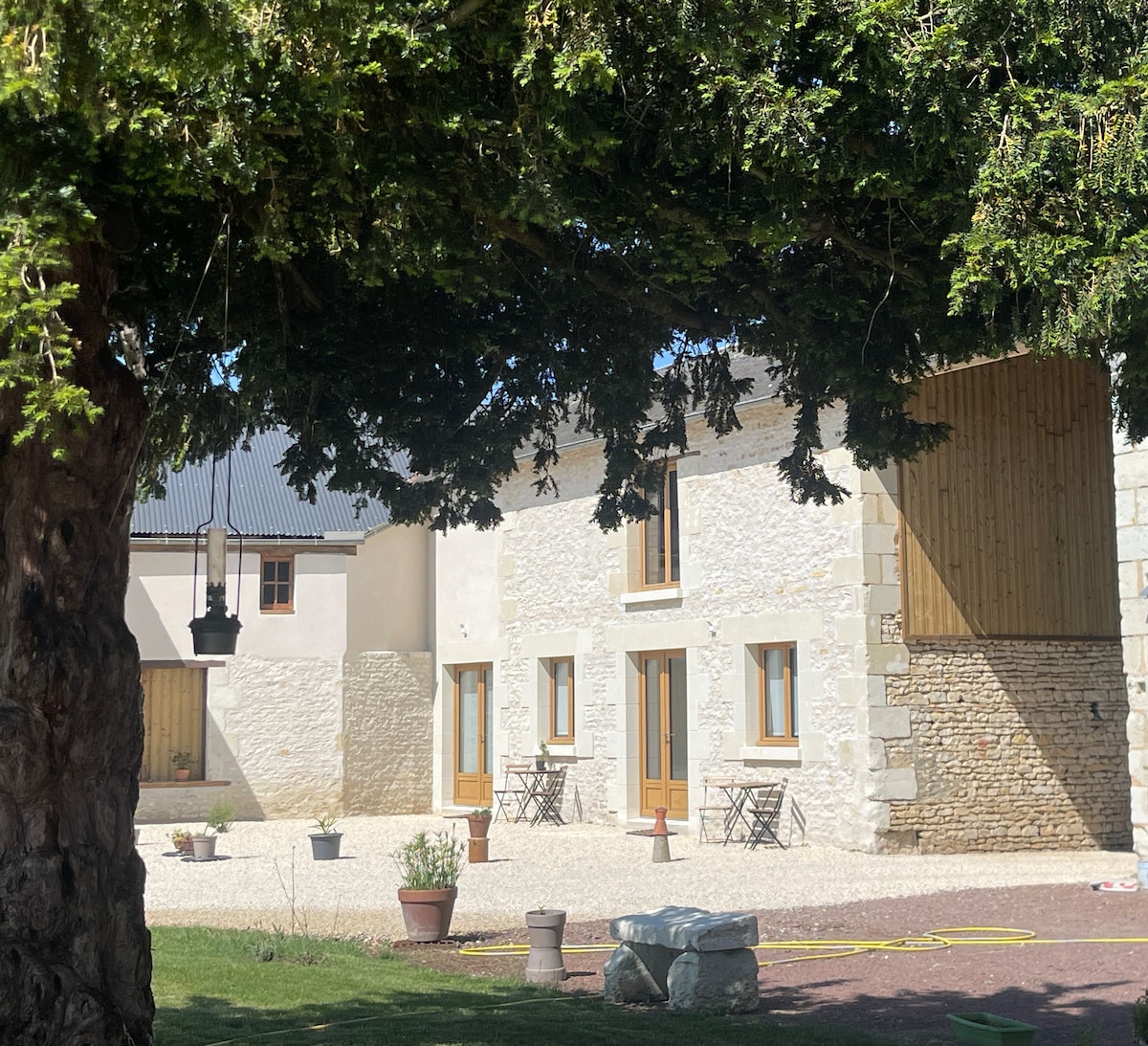 The exterior of a charming stone building is framed by lush greenery from a large tree. Small tables with chairs are situated on a gravel area in front of the building, which features multiple doors and windows, inviting natural light.