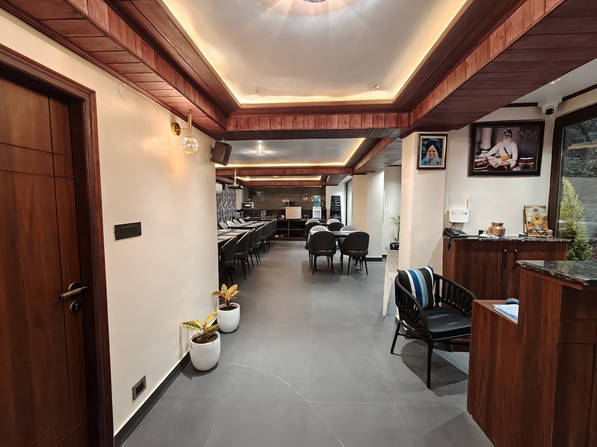 An inviting common area features sleek wooden accents and soft lighting. A central seating arrangement with dark chairs surrounds a wooden counter. Plants are placed in the corners, enhancing the contemporary decor. Large windows allow natural light to fill the space.