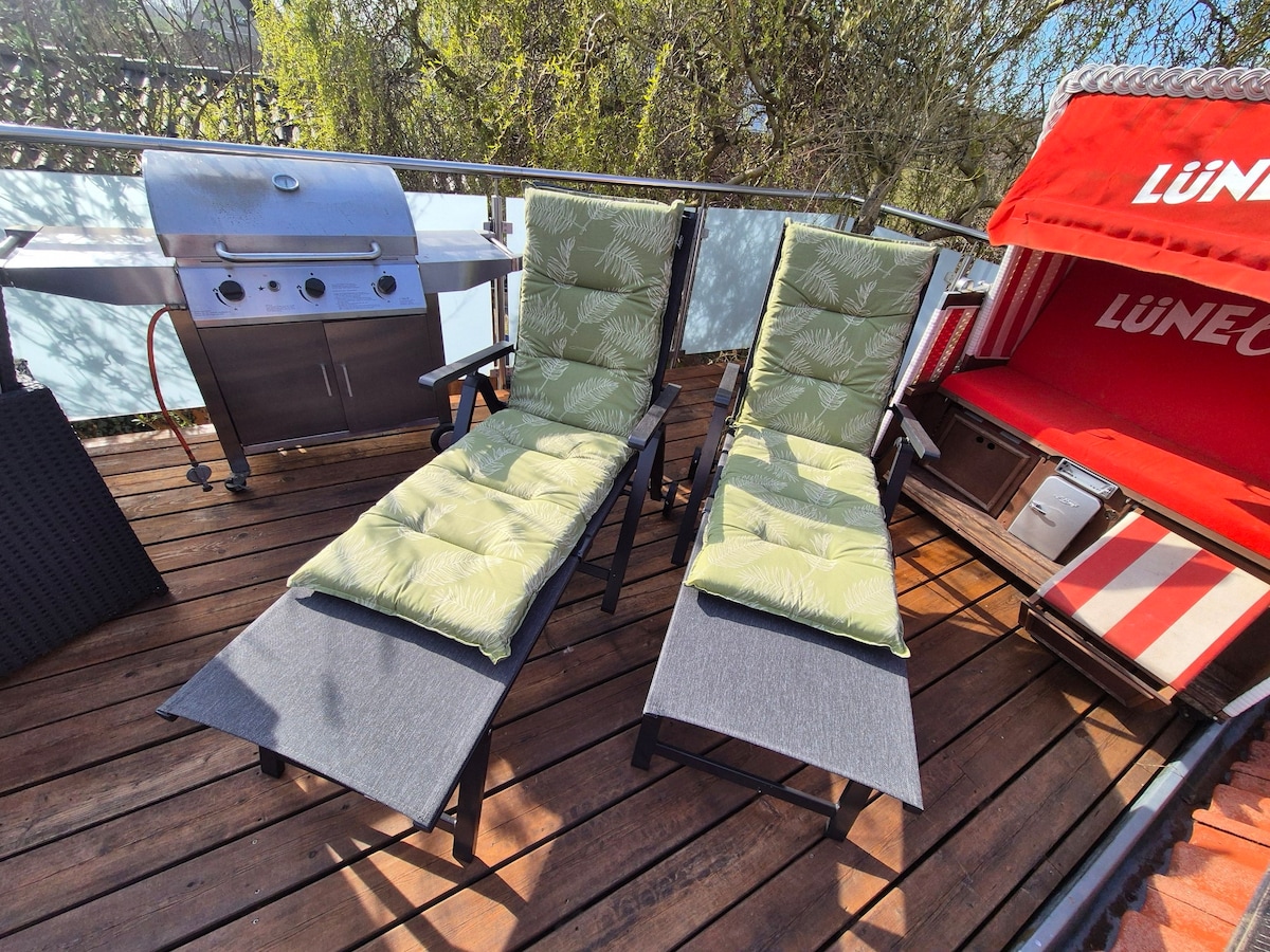Two comfortable lounge chairs with soft green cushions are positioned on a wooden deck. A gas grill is visible in the background, ready for outdoor cooking. A shaded beach chair adds a relaxing touch to the space.