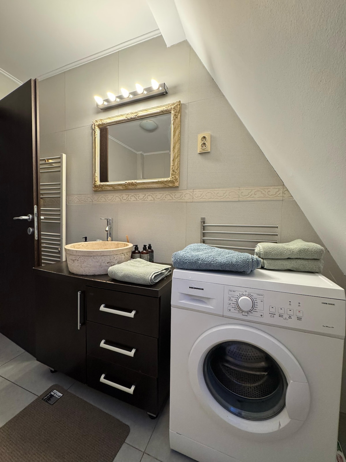 A well-appointed bathroom is visible, featuring modern fixtures and a large mirror. A washing machine sits beside a dark wood cabinet with drawers. Soft towels are neatly arranged on the countertop, while a woven basket adds texture to the space. A towel radiator is mounted on the wall.