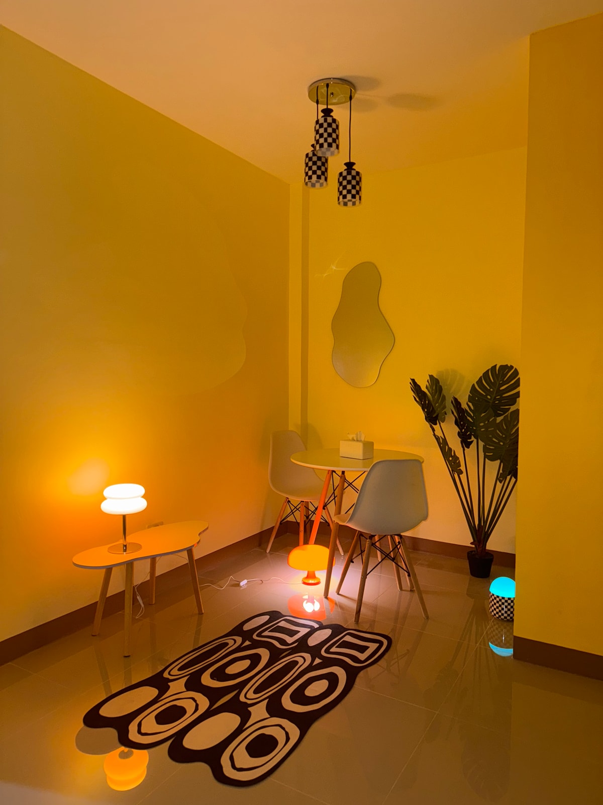 A cozy corner of the apartment features a small table with two light-colored chairs, complemented by a colorful rug on the tiled floor. A vibrant yellow wall adds warmth to the space, while soft lighting from several lamps creates a relaxing atmosphere. A potted plant adds a touch of nature.